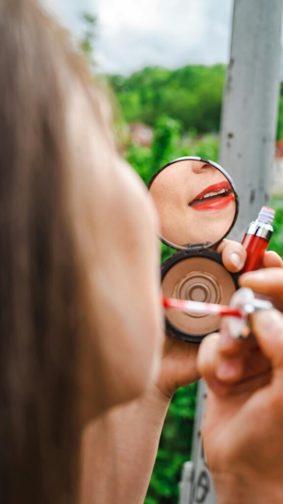Close-up of a woman applying red lipstick while looking into a compact mirror outdoors.